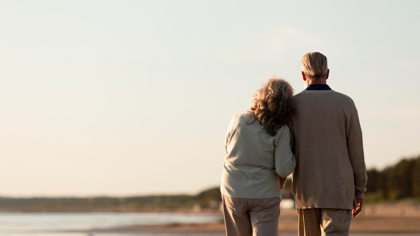 Una pareja de ancianos camina de la mano por la playa al atardecer. La mujer, de cabello rizado y canoso, se apoya en el hombro del hombre, quien lleva un suéter claro. Ambos lucen cómodos y felices en su paseo, con un paisaje de playa y un cielo suave de fondo, creando una atmósfera romántica y tranquila