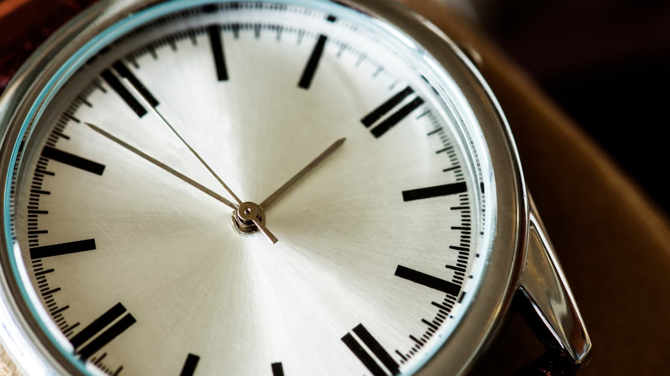 Un primer plano de la esfera de un reloj de pulsera muestra un diseño elegante con números en formato de línea negra y agujas en movimiento. La superficie de la esfera tiene un acabado brillante y metálico, mientras que el fondo desdibuja parcialmente el cierre de la correa, sugiriendo un accesorio de moda atemporal