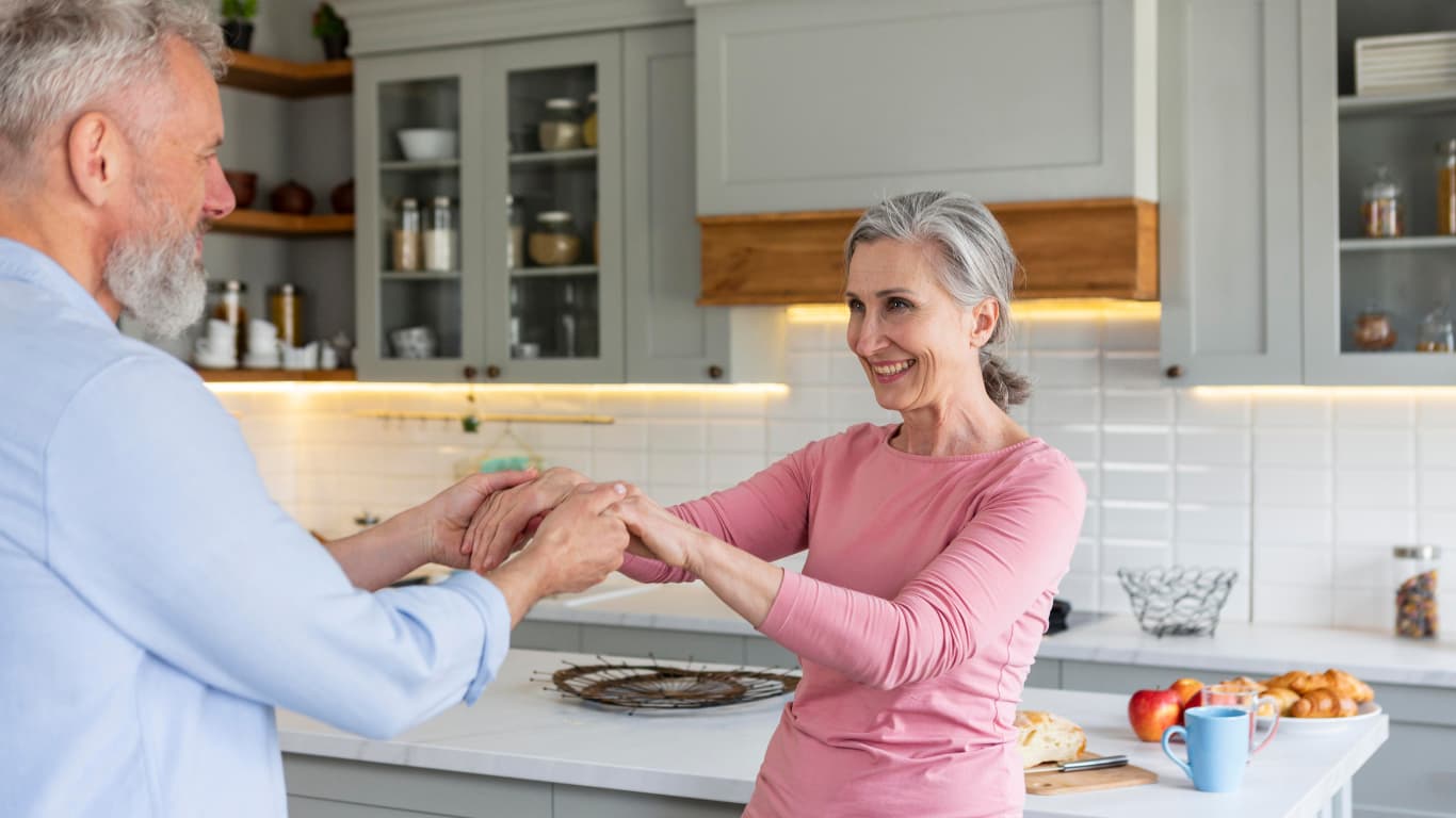 Una pareja de adultos mayores se ríe y se toma de las manos en una cocina moderna y luminosa. El hombre, con cabello y barba canosa, lleva una camisa de mezclilla, mientras que la mujer, con cabello plateado, viste una blusa rosa.
