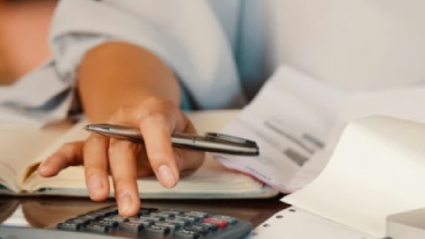 Una mano sostiene un bolígrafo mientras presiona un botón en una calculadora. En la mesa hay un cuaderno abierto, así como papeles desordenados, sugiriendo un ambiente de trabajo en el que se están realizando cálculos o tomando notas
