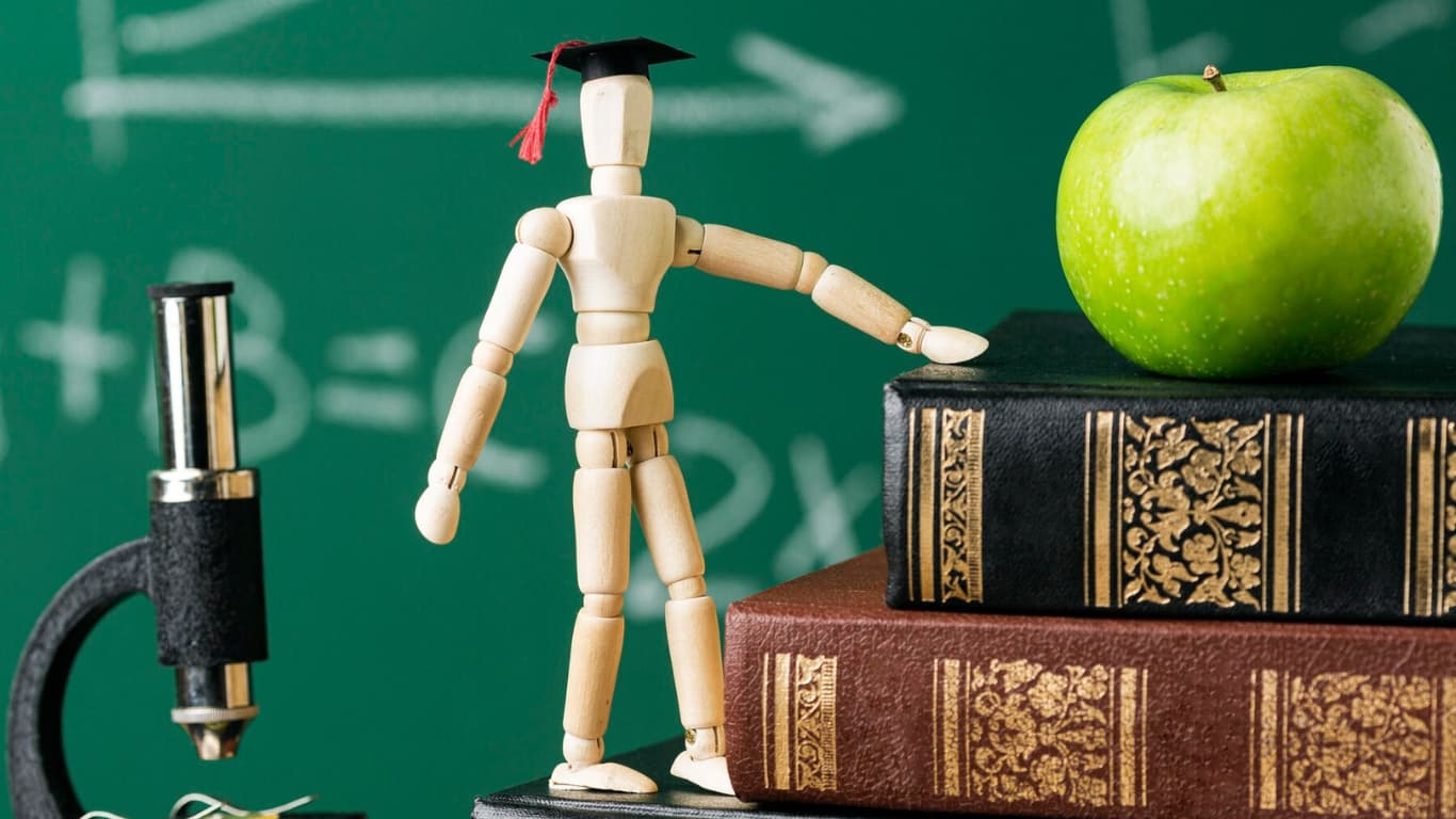 Representación de la educación con maniquí de graduación, libros antiguos, microscopio y manzana verde frente a un pizarrón.