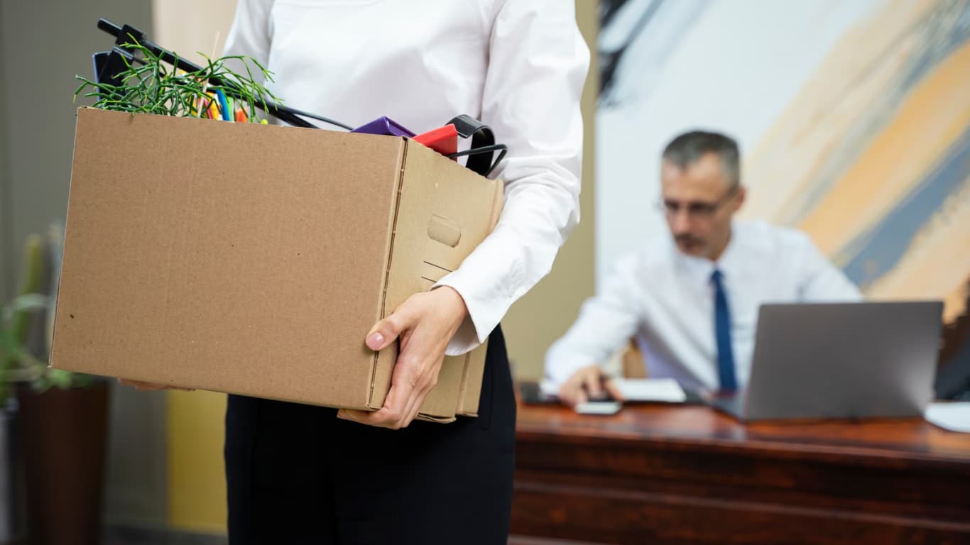 Una mujer sostiene una caja de cartón llena de suministros de oficina, mientras camina por una oficina.