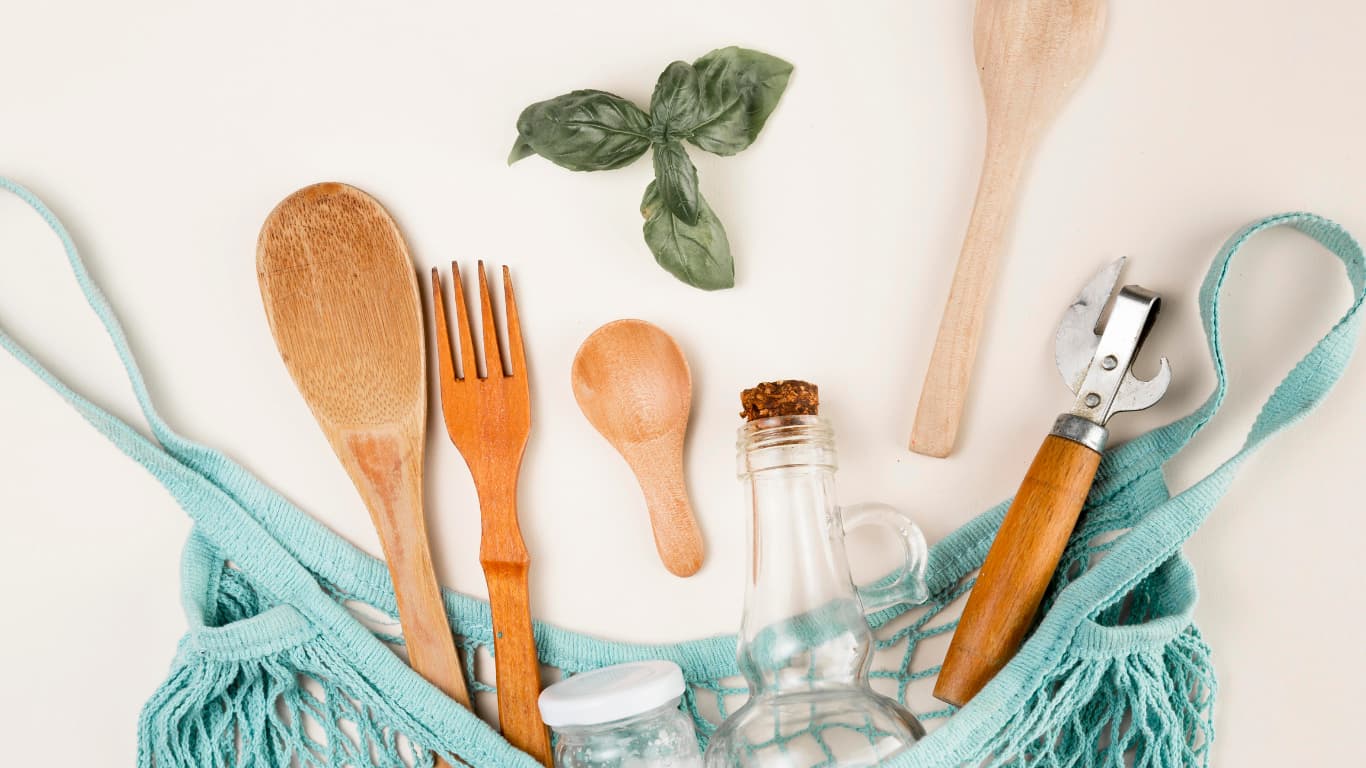 Una variedad de utensilios de cocina dispuestos sobre un fondo claro. Se incluyen cucharas de madera, un tenedor, un sacacorchos, y frascos de vidrio, junto con hojas de albahaca fresca.