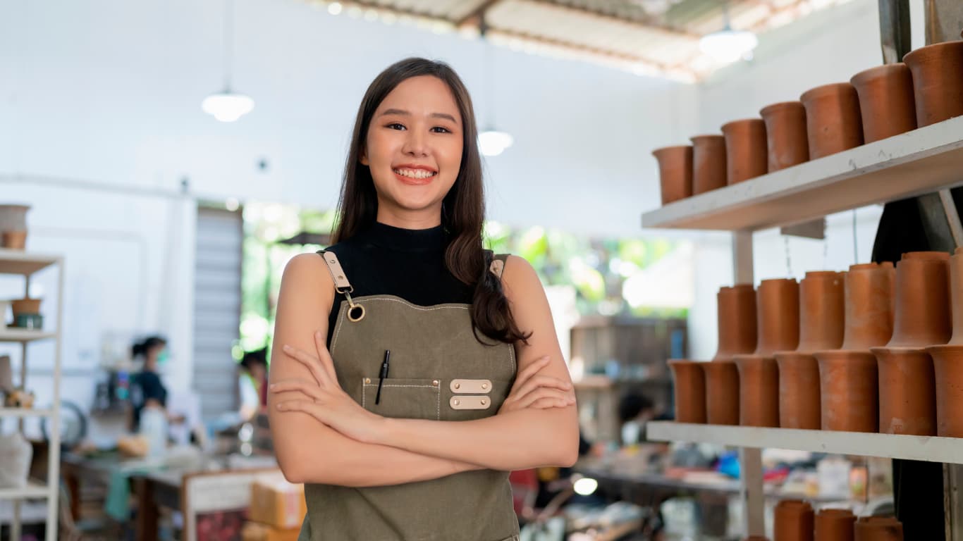 Una joven sonriente está de pie en un taller, con los brazos cruzados y un delantal verde. Ella muestra confianza y alegría, mientras el fondo presenta estantes con macetas de barro y un ambiente luminoso y creativo.