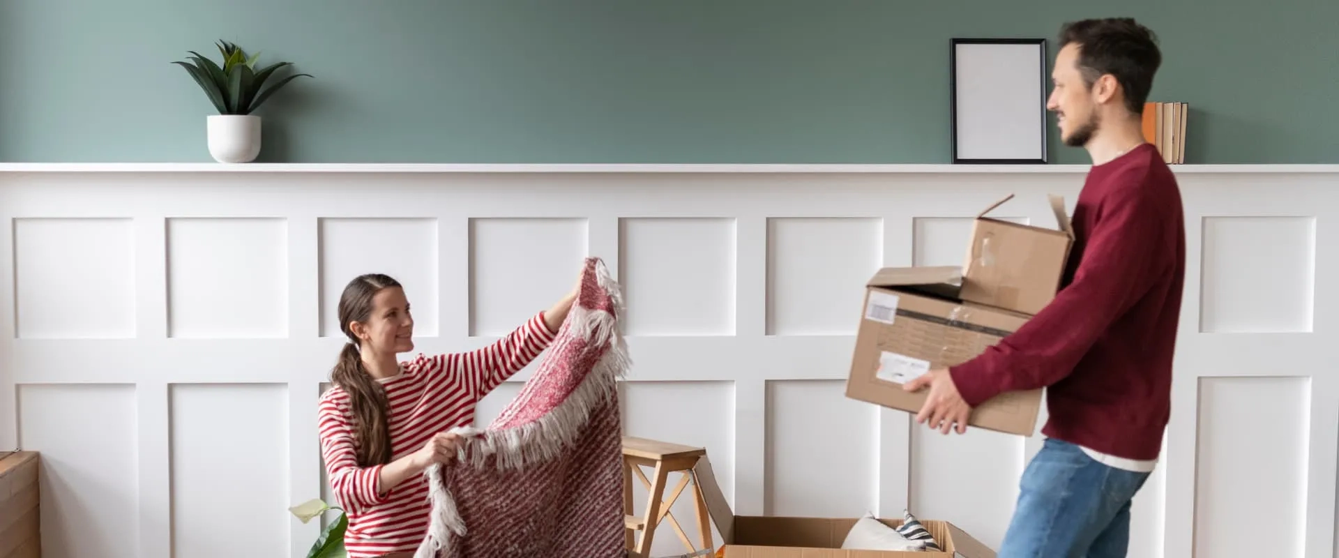 Una mujer sentada en el suelo sostiene una manta mientras mira a un hombre que lleva cajas de cartón. Ambos están en un espacio interior con paredes verdes y decoraciones minimalistas. Hay varias cajas y un taburete en el entorno, sugiriendo una mudanza o reordenamiento.