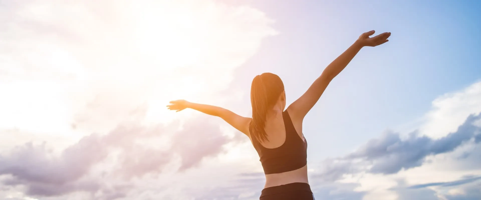 Una mujer de espaldas con los brazos extendidos hacia el cielo, disfrutando de la luz del sol. Ella viste una camiseta negra y tiene el cabello amarrado en una coleta. El fondo muestra un cielo parcialmente nublado con rayos de sol brillando, creando una atmósfera de libertad y bienestar