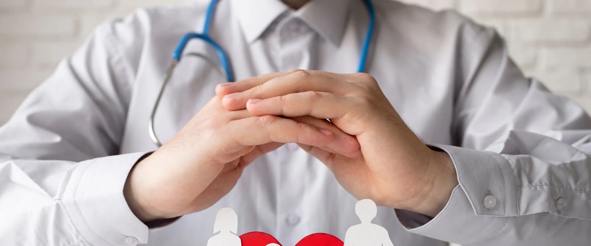 Un profesional de la salud con una camisa blanca y un estetoscopio azul alrededor del cuello coloca sus manos en forma de triángulo frente a una ilustración de figuras humanas y un corazón rojo en la parte inferior. El fondo presenta una pared de ladrillos claros, creando un ambiente profesional y acogedor