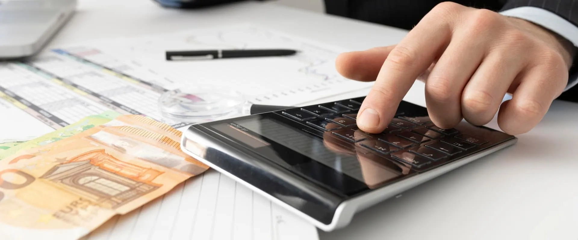 Un primer plano de una mano masculina usando una calculadora sobre una mesa con documentos y billetes de euro. En el fondo, se puede ver una hoja de cálculo con gráficos y un bolígrafo, sugiriendo una actividad relacionada con finanzas o contabilidad.