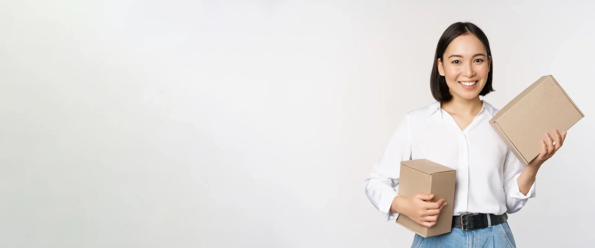 Una mujer joven con cabello corto y oscuro sonríe mientras sostiene dos cajas de cartón en un fondo claro. Lleva una camisa blanca y pantalones de cintura alta. Su expresión transmite alegría y satisfacción, sugiriendo un ambiente relacionado con el envío o la entrega de paquetes