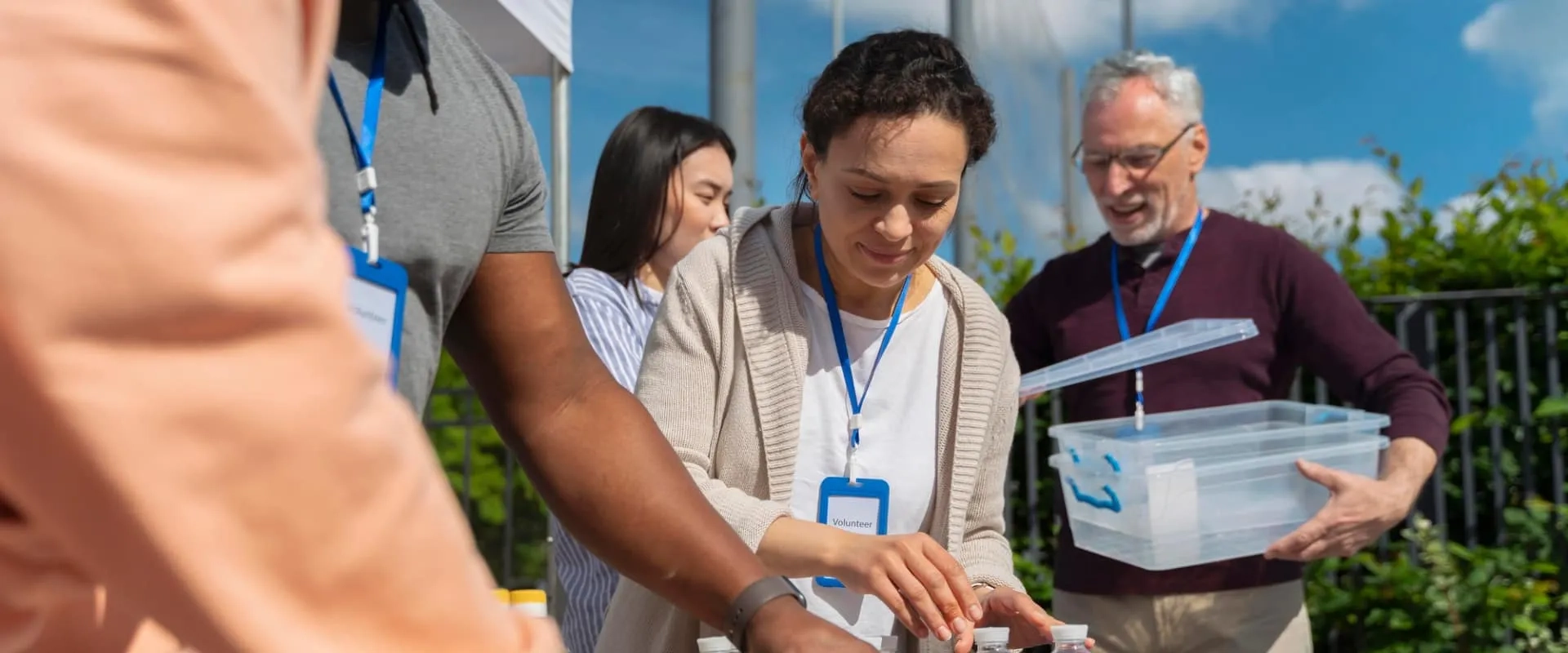 Un grupo de cuatro voluntarios trabaja al aire libre en un evento comunitario. En el primer plano, una mujer sonriente con una etiqueta de 'voluntario' está organizando algunos elementos sobre una mesa, mientras que un hombre a su lado sostiene una caja de almacenamiento transparente.