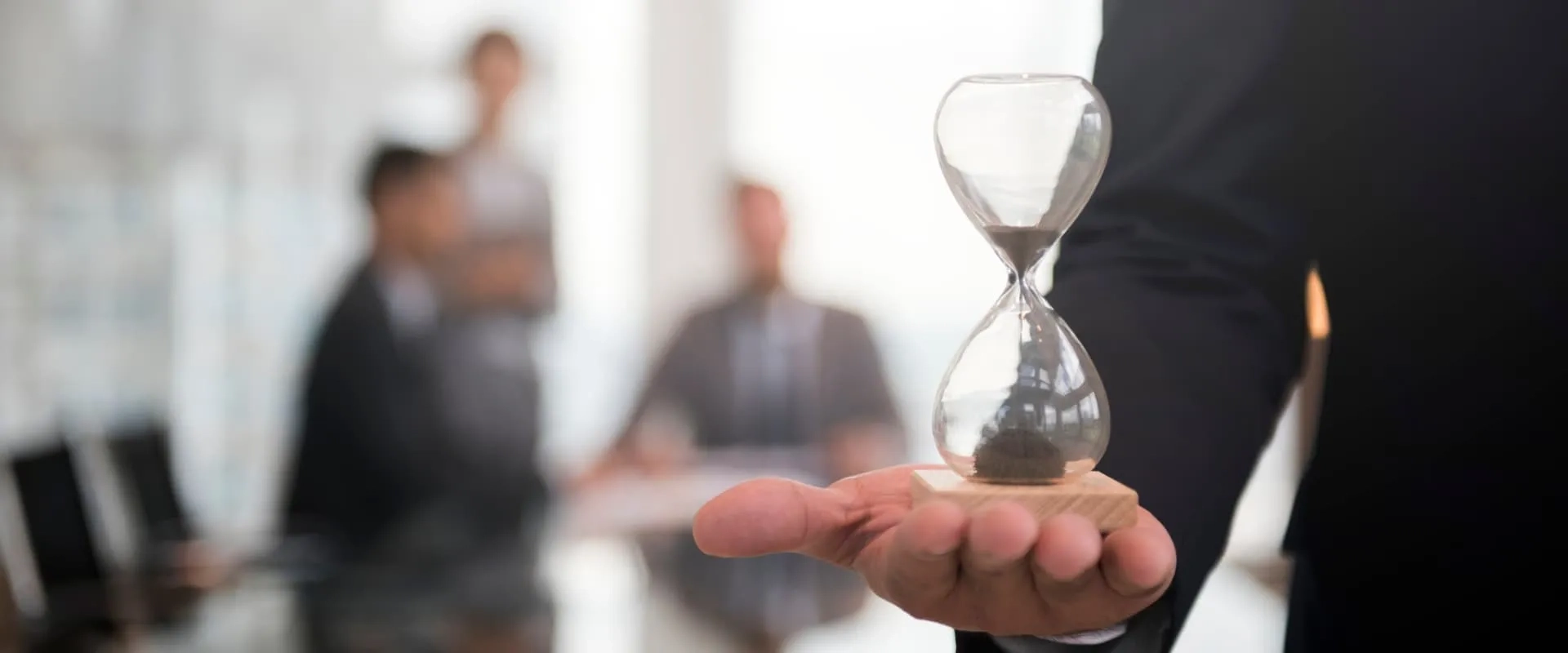 Un hombre con traje sostiene un reloj de arena sobre su mano abierta en un entorno de oficina. Al fondo, se pueden ver siluetas de varias personas en una reunión. La imagen evoca conceptos de gestión del tiempo y toma de decisiones en un contexto profesional