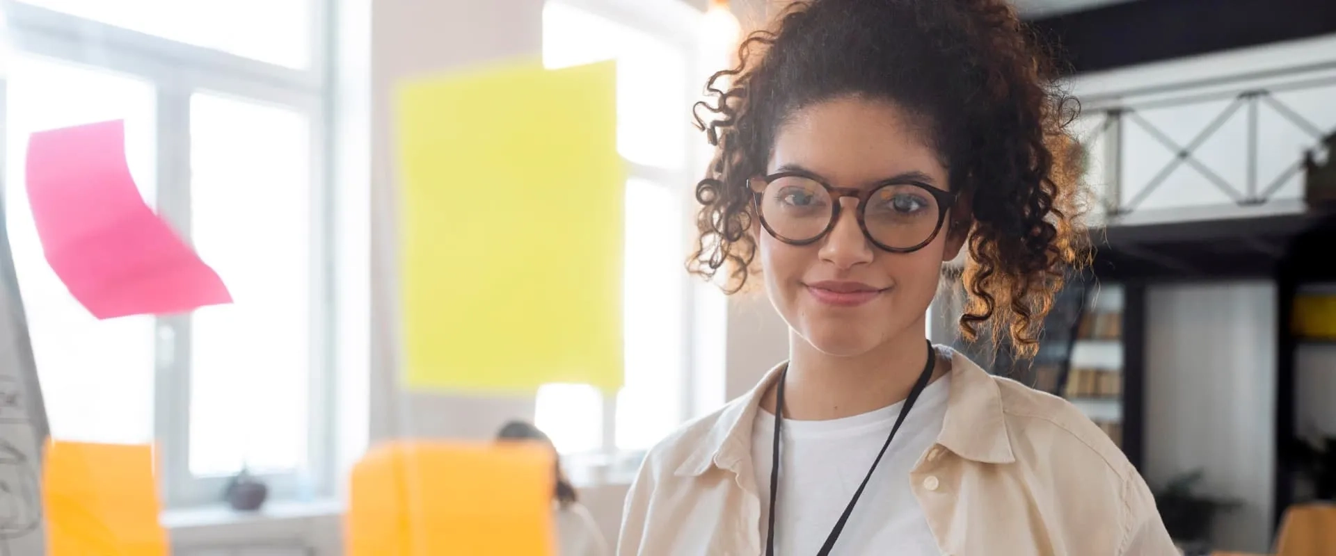 Una mujer sonriente con cabello rizado y gafas de gran tamaño sostiene un bloc de notas rosa en una mano y un marcador en la otra. Está en un ambiente de oficina moderno, con notas adhesivas de varios colores visibles en el fondo y gente trabajando sentada en mesas. La luz natural entra por las ventanas, creando un ambiente acogedor y creativo