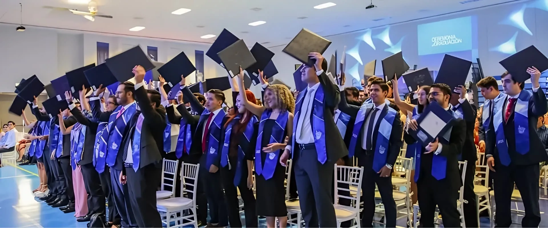 Un grupo de graduados en una ceremonia levantan sus diplomas en alto en señal de celebración. Todos visten trajes formales y pañuelos de graduación en tonos azules. El ambiente es festivo mientras los graduados sonríen y celebran sus logros, y se pueden ver asientos vacíos en el fondo