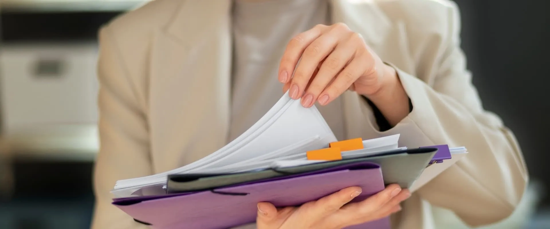 Una persona sostiene una carpeta con documentos en una mano y revisa unas hojas con la otra. La persona lleva puesto un traje claro y el fondo es desenfocado, sugiriendo un ambiente de oficina.