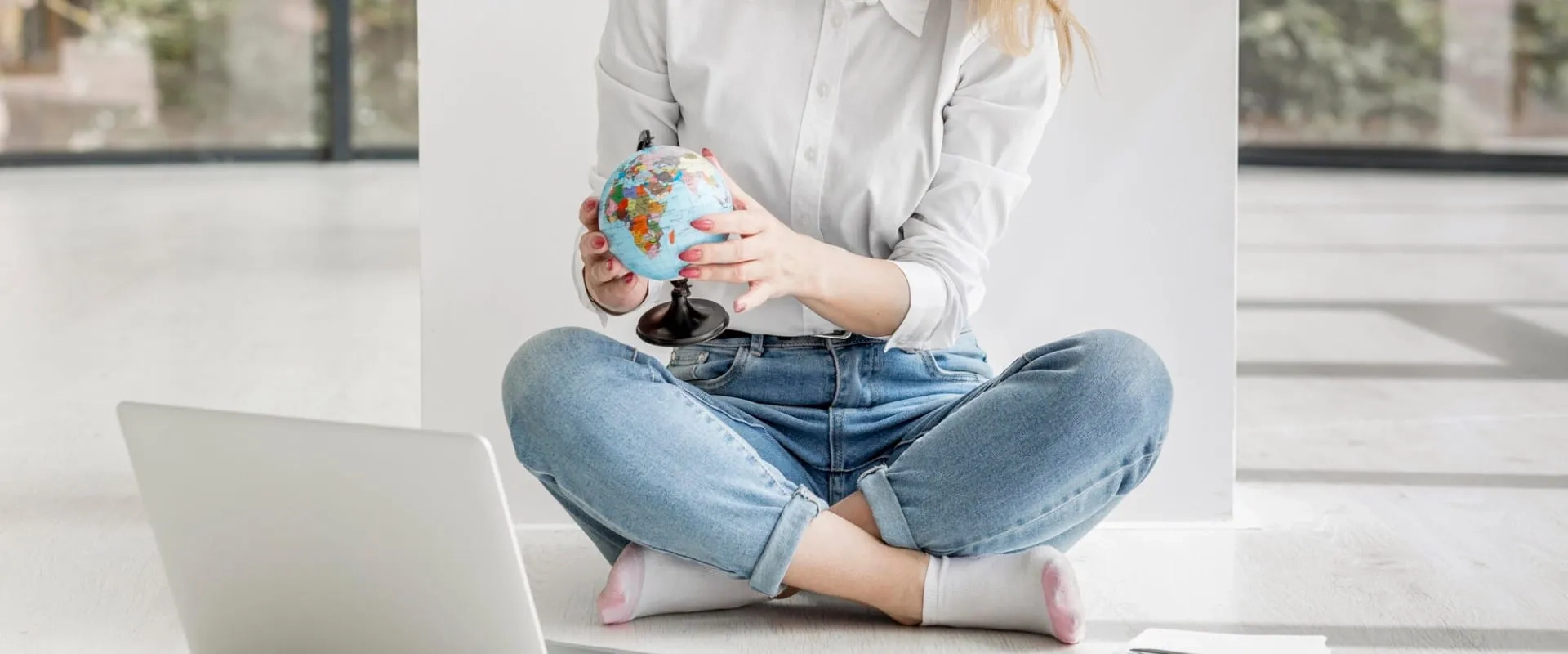 Mujer joven sentada en el suelo con un globo terráqueo y una laptop, planificando estudios o viajes internacionales.