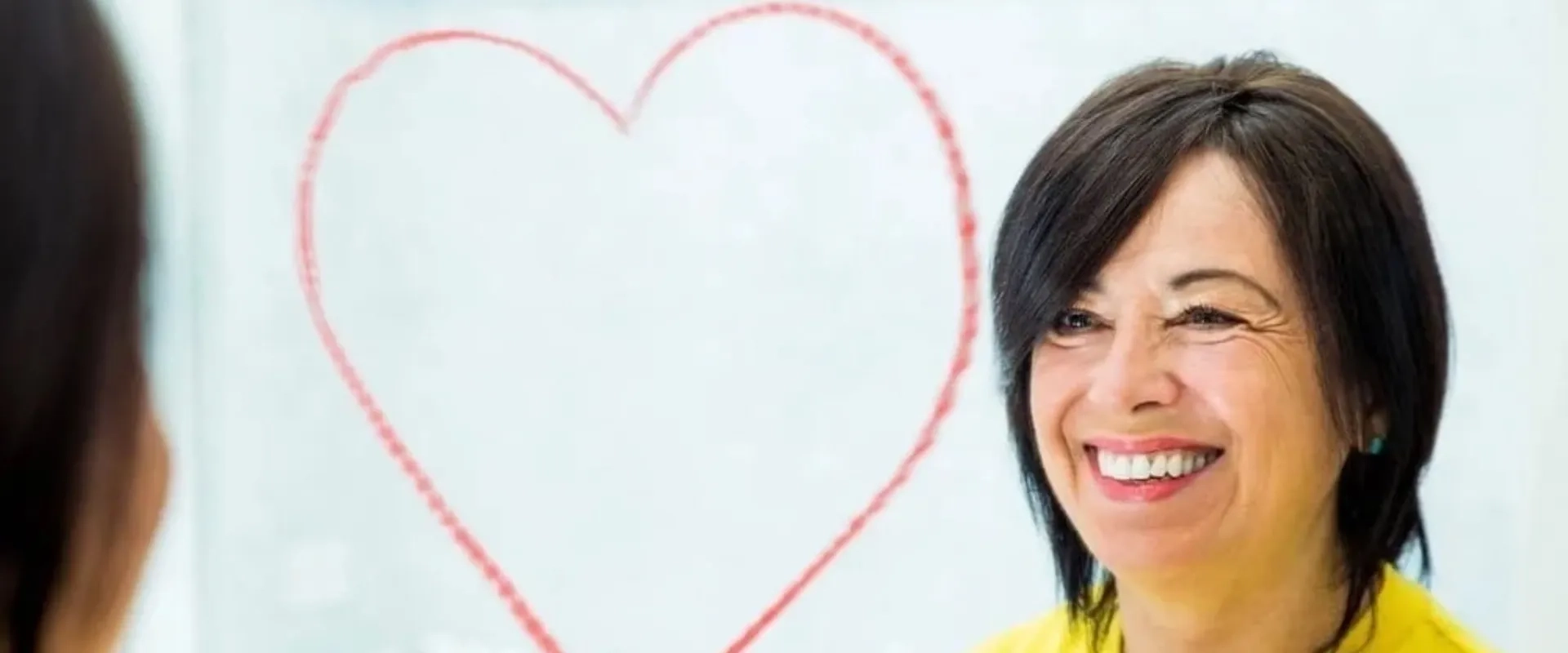 Una mujer sonriente con cabello corto y oscuro, mirando hacia un espejo. Detrás de ella, hay un dibujo de un corazón en la pared, lo que sugiere un ambiente positivo y afectuoso.