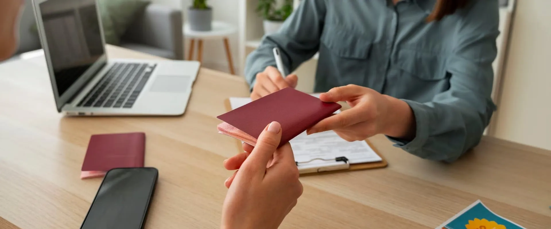 Una persona entregando un pasaporte a otra en una mesa de trabajo. En el fondo, hay una laptop, un cuaderno con notas y un teléfono móvil. 