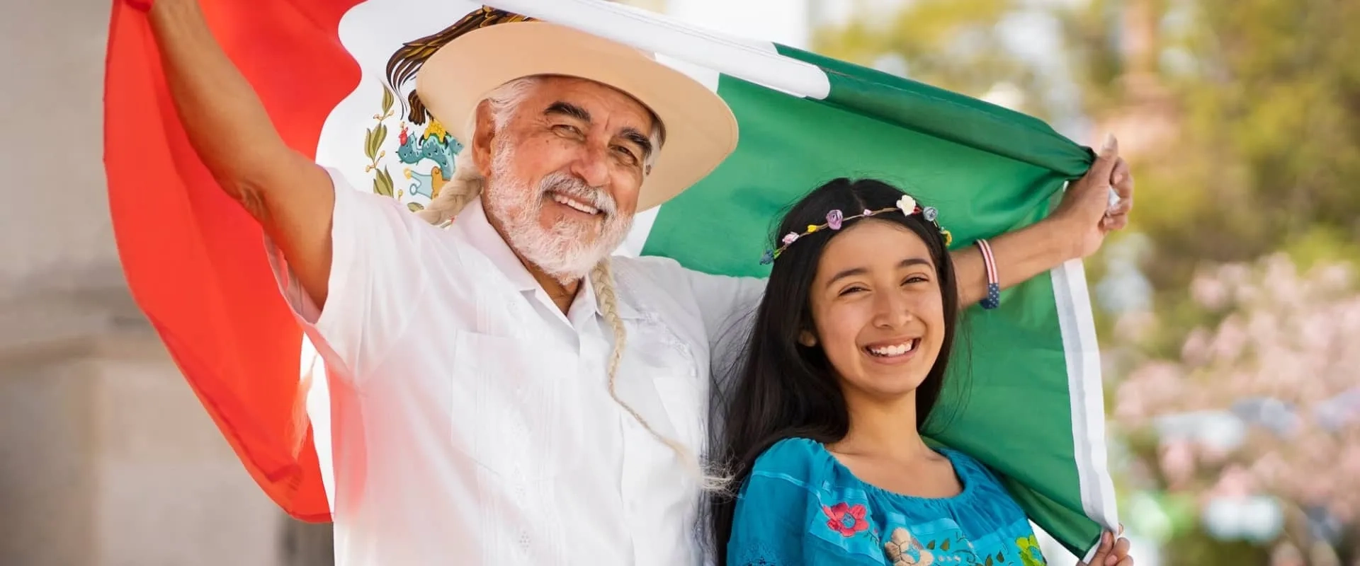 Un hombre mayor con barba y un sombrero está sonriendo mientras sostiene una bandera de México junto a una niña que lleva un vestido tradicional.