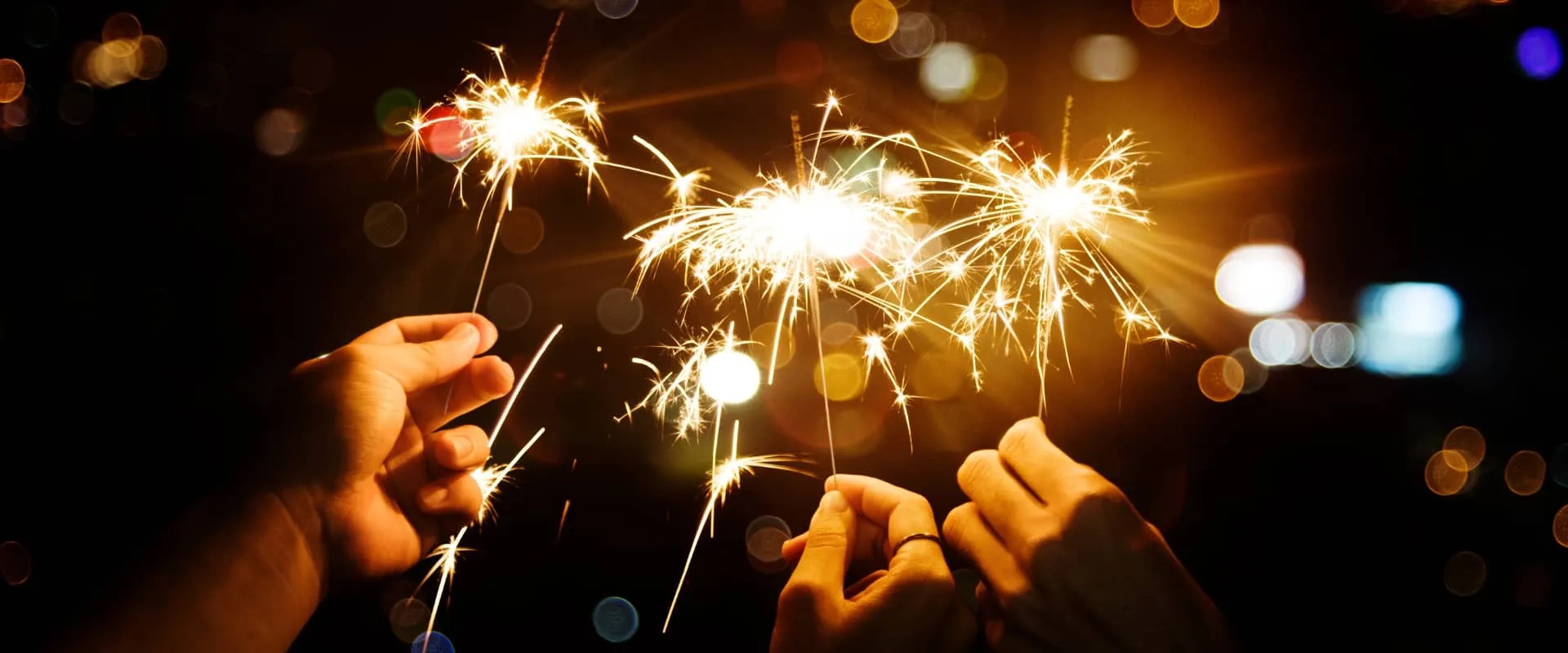 Varias manos sostienen varitas de chispa encendidas, creando destellos de luz sobre un fondo oscuro. Los destellos brillantes iluminan la escena, mientras se pueden ver luces borrosas en el fondo, sugiriendo una celebración o un momento festivo.