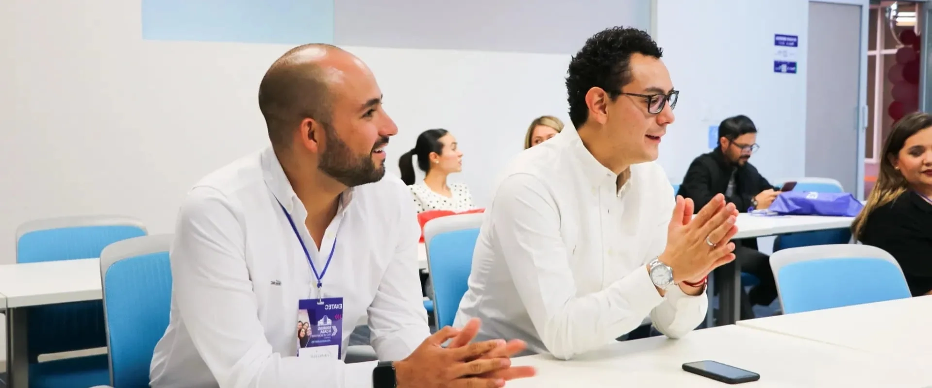 Dos hombres sentados en una sala de reuniones, ambos vistiendo camisas blancas y sonriendo. Están atentos, con las manos juntas en un gesto de interés. En el fondo, se pueden ver otras personas participando en la reunión.