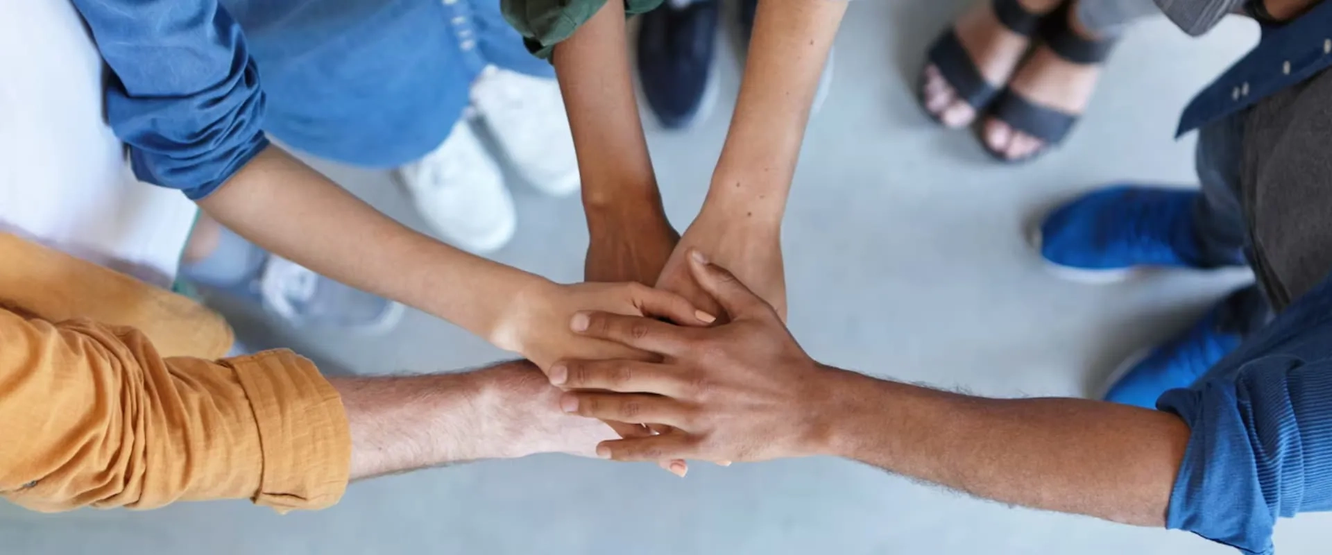 Manos de diferentes personas están apiladas unas sobre otras en una mesa. La imagen captura un momento de cooperación y apoyo, con un fondo desenfocado que muestra el suelo. 