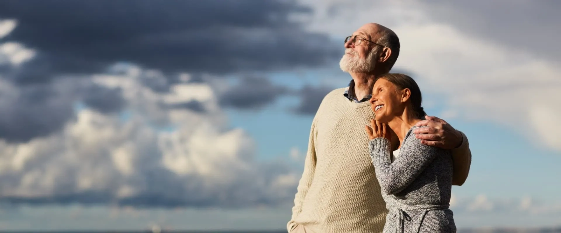 Una pareja de personas mayores está de pie al aire libre, abrazados y mirando hacia el cielo. El hombre tiene barba y gafas, mientras que la mujer sonríe con alegría. 