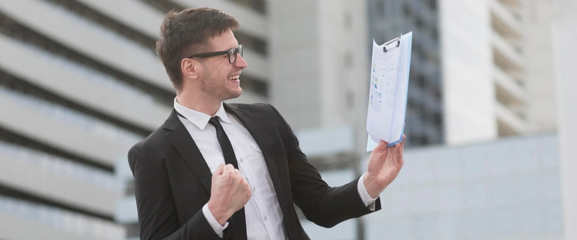 Un hombre vestido con un traje negro y gafas, sonriendo y levantando una carpeta con documentos en una mano.
