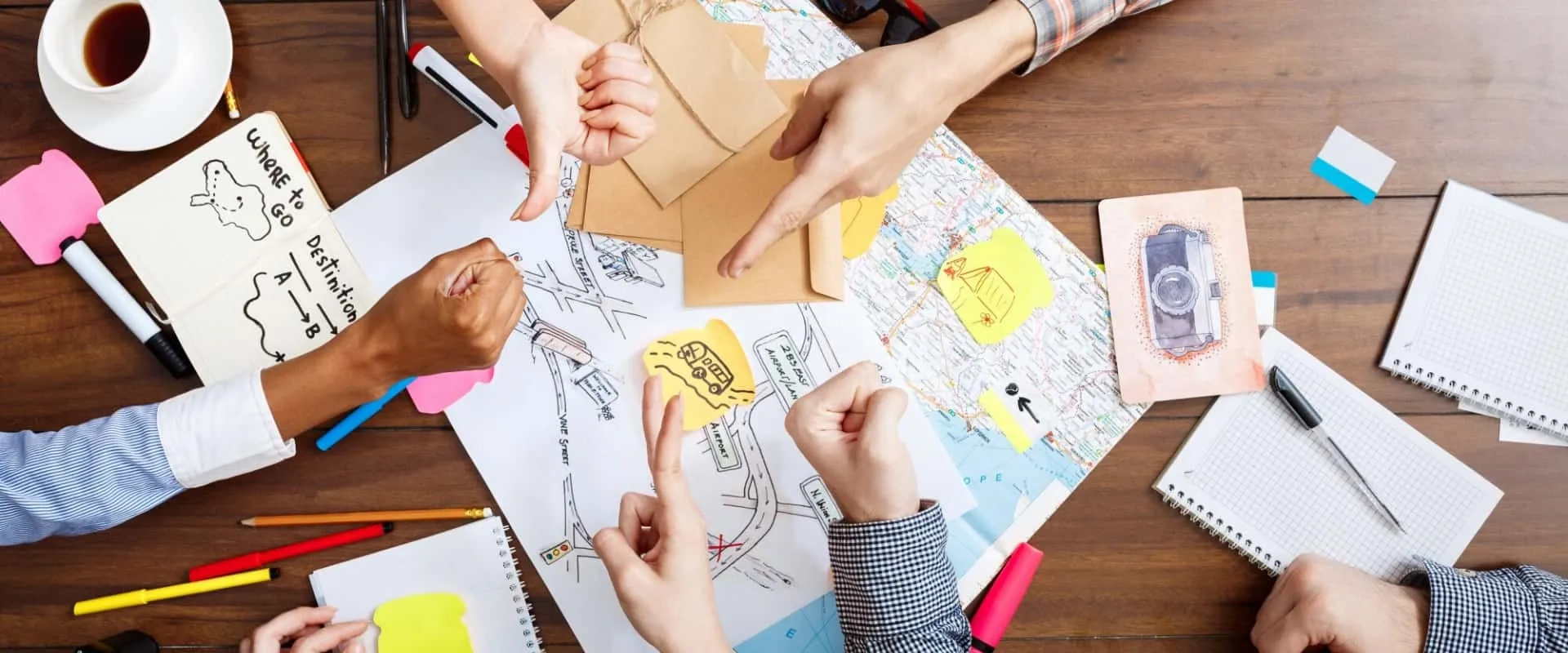 Varios brazos de personas colaborando en una mesa, señalando un mapa y notas adhesivas de colores. 