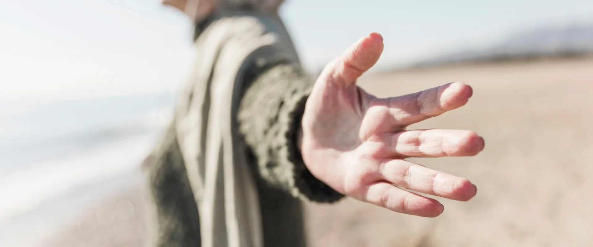 Un primer plano de una mano extendida hacia el espectador, con un fondo de playa y mar desenfocado.