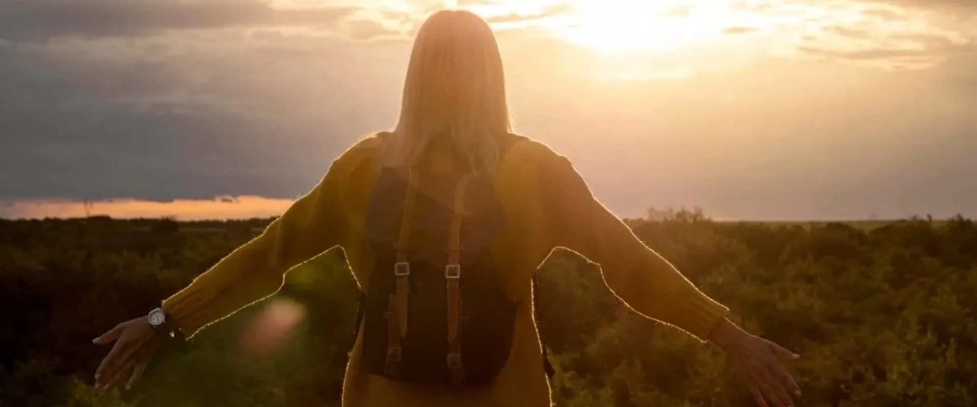Una mujer de espaldas, con un suéter amarillo y una mochila, de pie frente a un paisaje natural bajo la luz del atardecer.
