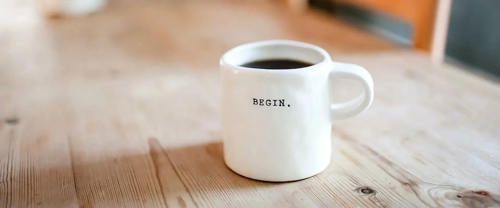 Una taza blanca con la palabra 'BEGIN.' impresa en negro está sobre una mesa de madera. La taza contiene café y la composición sugiere un ambiente acogedor y motivador, ideal para empezar el día con energía.