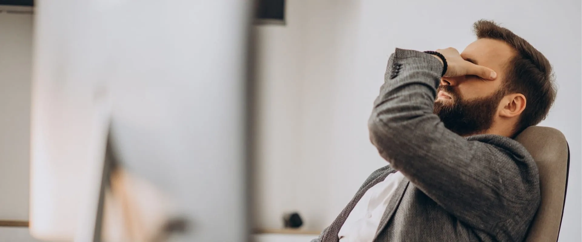 Un hombre con barba y cabello corto está sentado en una silla frente a una computadora, con una mano en su frente, mostrando signos de estrés o frustración. Estrés laboral. 