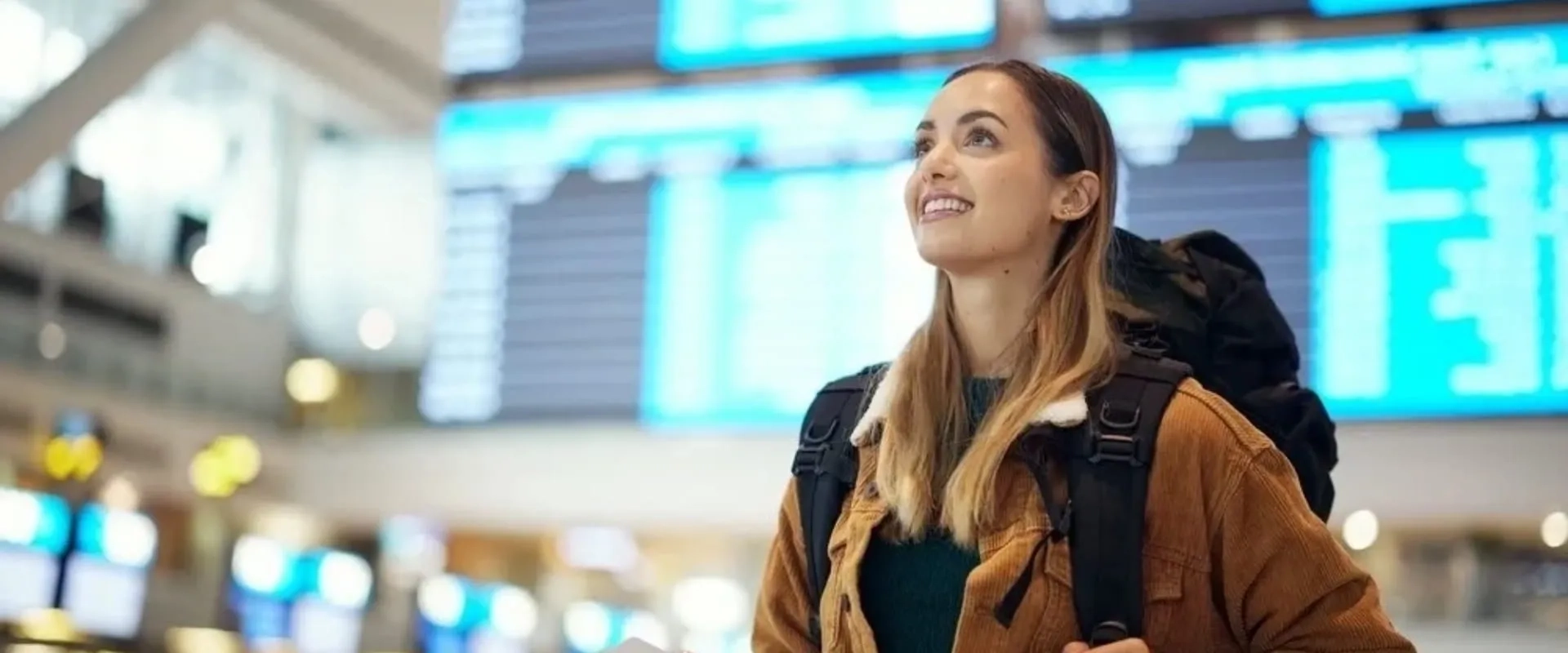 Una joven sonriente con una mochila grande está en un aeropuerto, mirando hacia las pantallas de llegadas y salidas. 