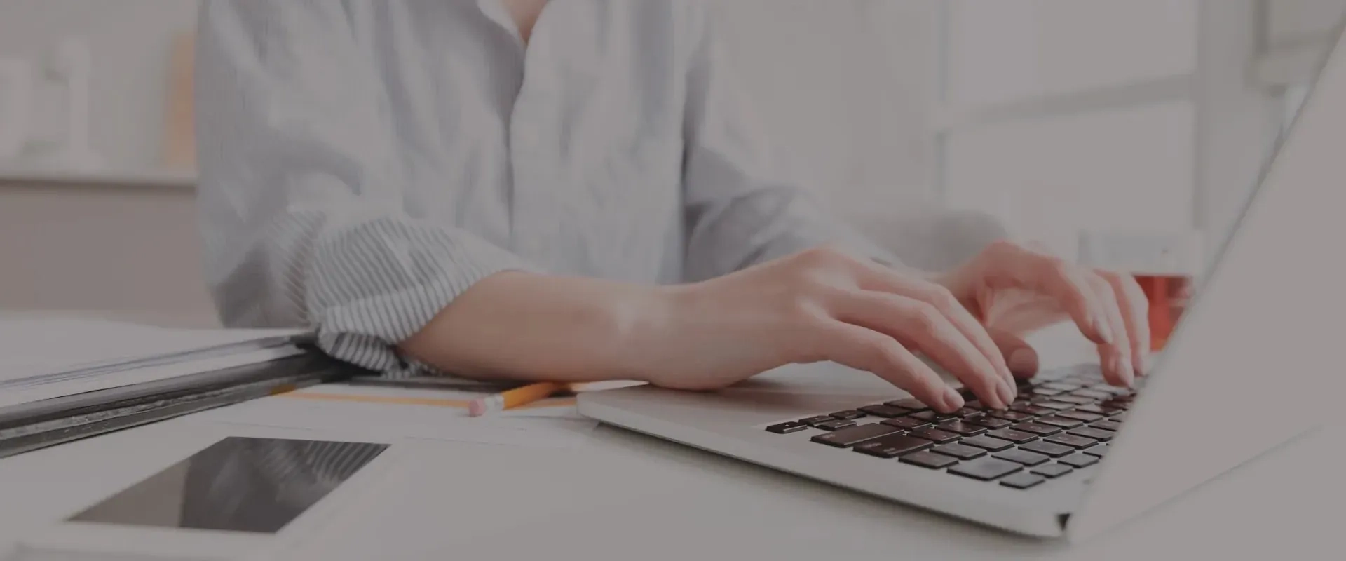 Primer plano de una persona tecleando en un ordenador portátil. La mano está sobre el teclado y se pueden ver algunos objetos de oficina en la mesa, como un teléfono inteligente, un cuaderno y un lápiz. 