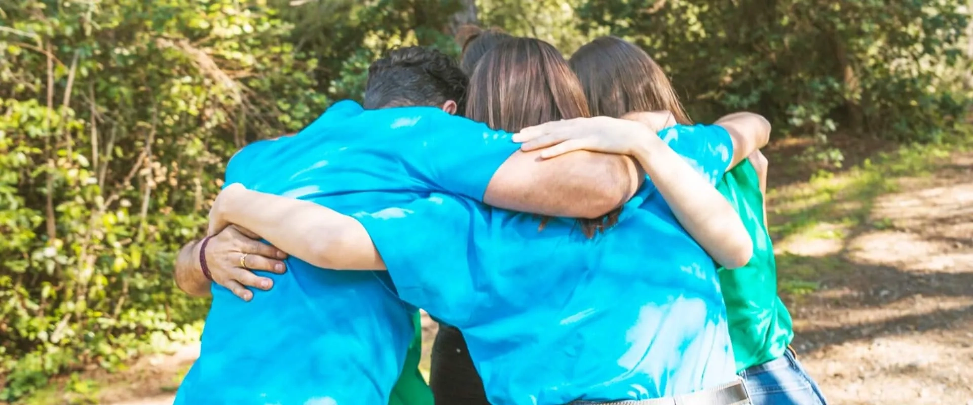 Un grupo de cuatro personas se abraza en un entorno natural. Todos llevan camisetas de colores brillantes, como azul y verde.