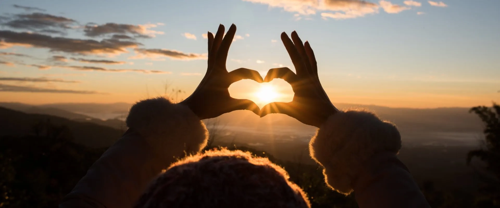 Una persona al aire libre forma un corazón con sus manos, enmarcando el sol que se eleva en el horizonte durante el amanecer.