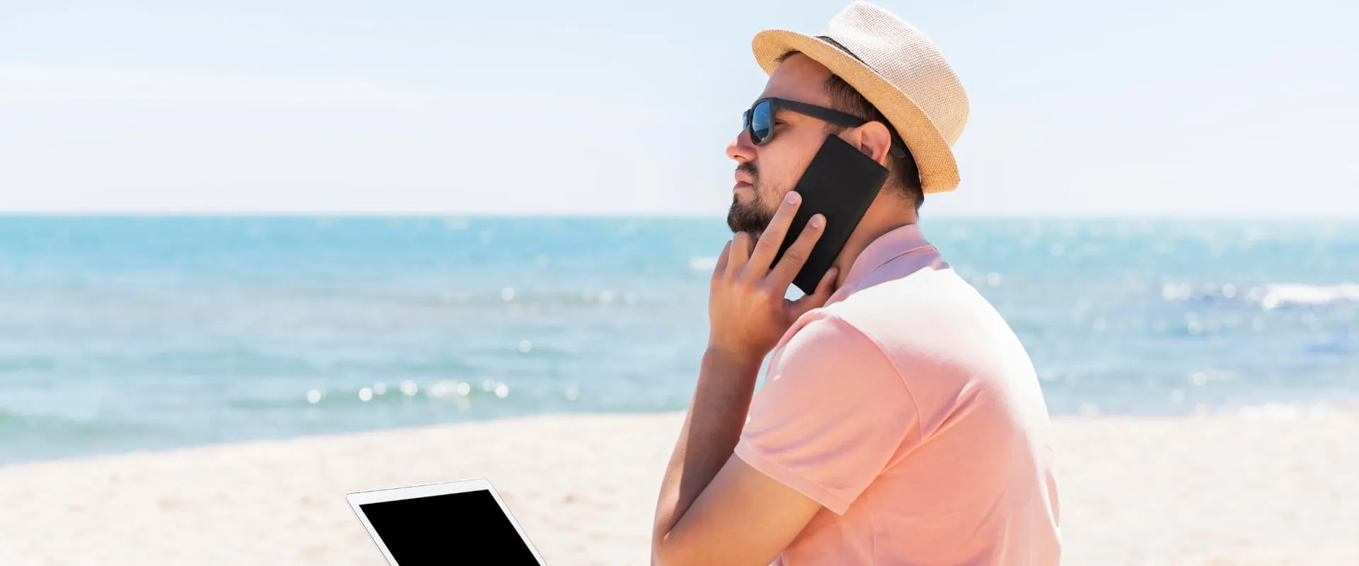 Un hombre con gafas de sol y un sombrero se encuentra en la playa, hablando por teléfono móvil.
