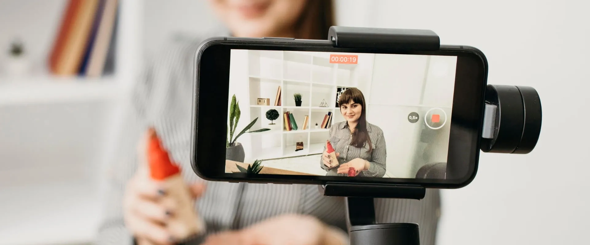 Una mujer sonriente está siendo grabada con un teléfono móvil montado en un trípode, mientras sostiene un envase rojo. La pantalla del teléfono muestra su imagen en un entorno de oficina, con estanterías decoradas al fondo. 