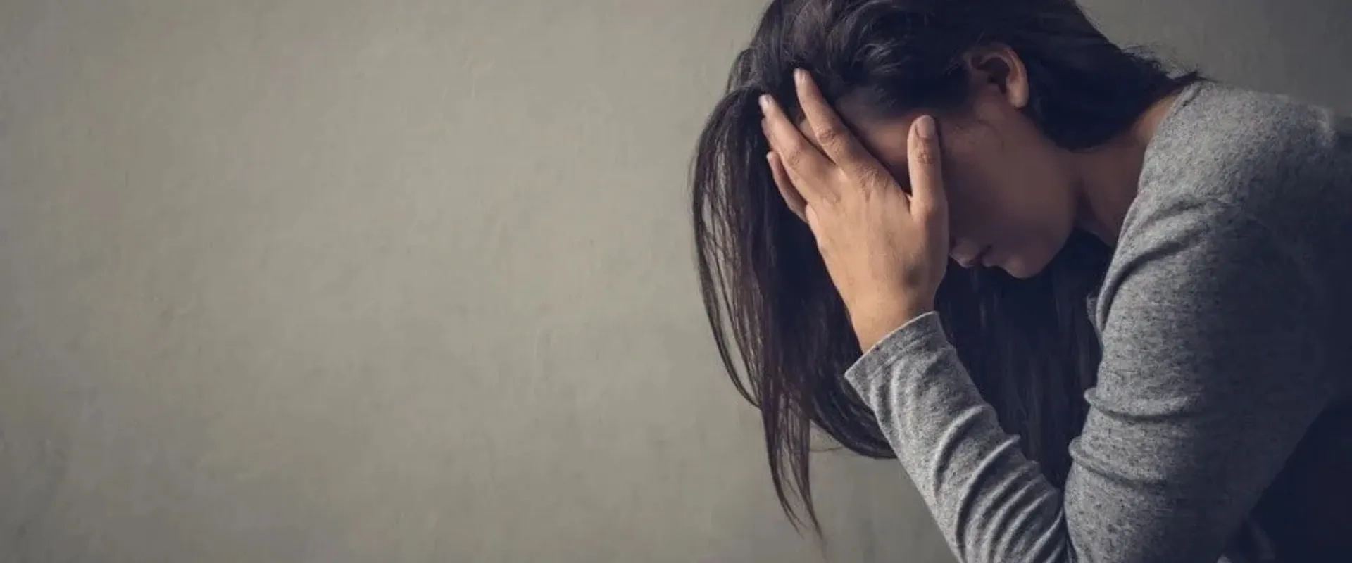 Una mujer con el cabello largo y oscuro se sienta con la cabeza agachada y las manos en la frente, mostrando una expresión de angustia o estrés.