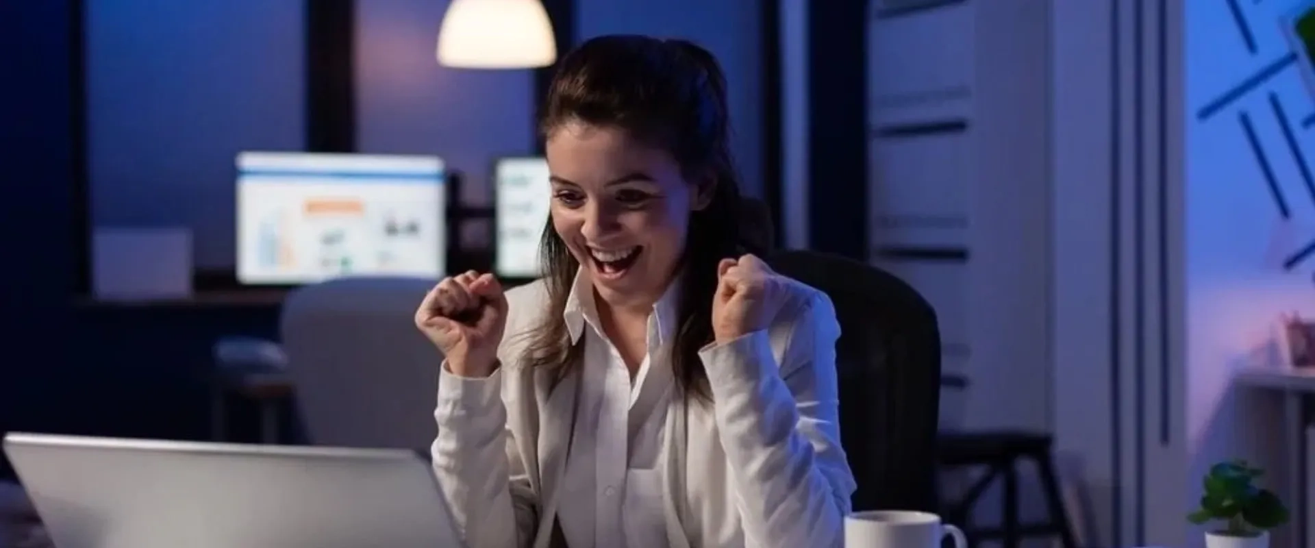Una mujer sonriente con cabello oscuro y suelto está sentada frente a una computadora portátil en un ambiente iluminado suavemente.