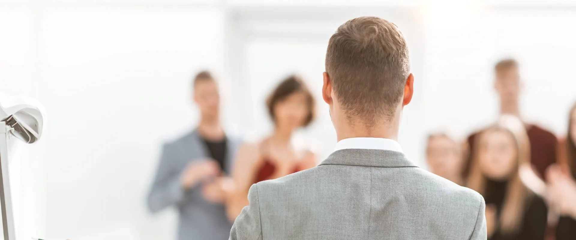 Un hombre de espaldas, vestido con un traje gris, se encuentra frente a un grupo de personas que aplauden. 