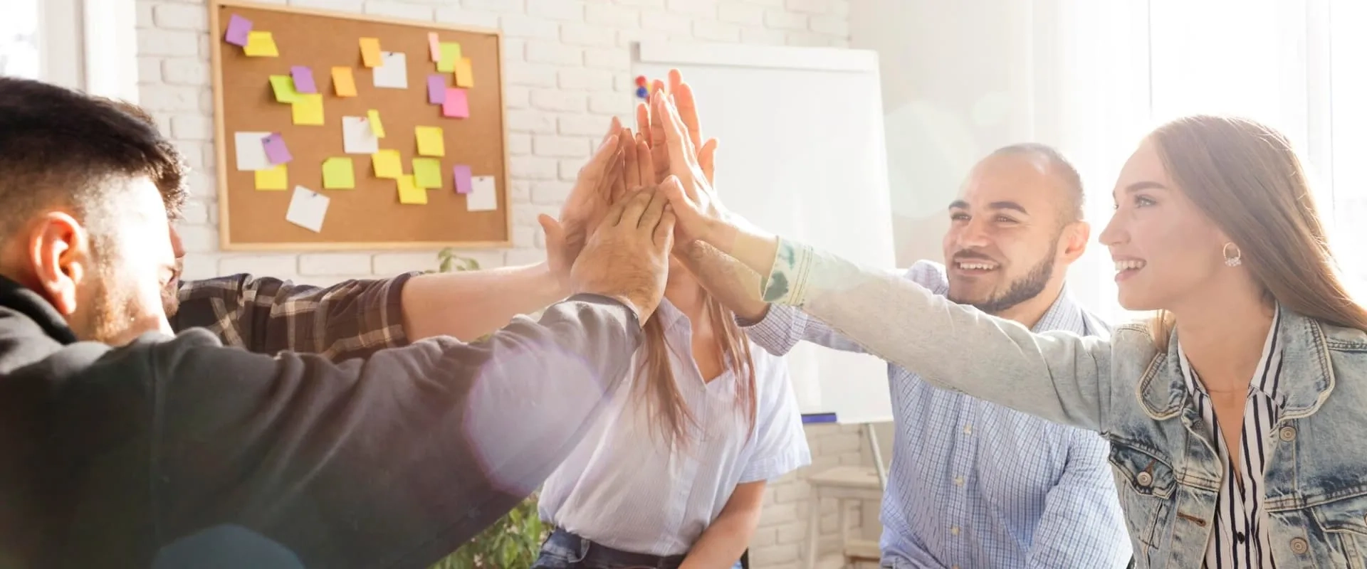 Un grupo de cuatro personas se da un 'high five' en un entorno de oficina iluminado, con una pizarra y notas de colores en el fondo.