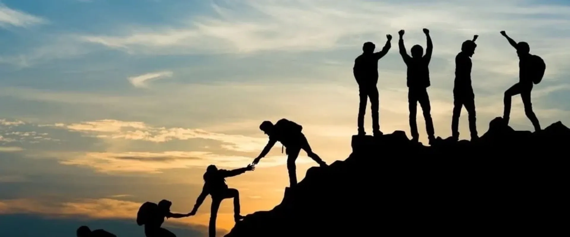 Siluetas de un grupo de personas en la cima de una montaña al atardecer, algunas levantando los brazos en señal de triunfo. 
