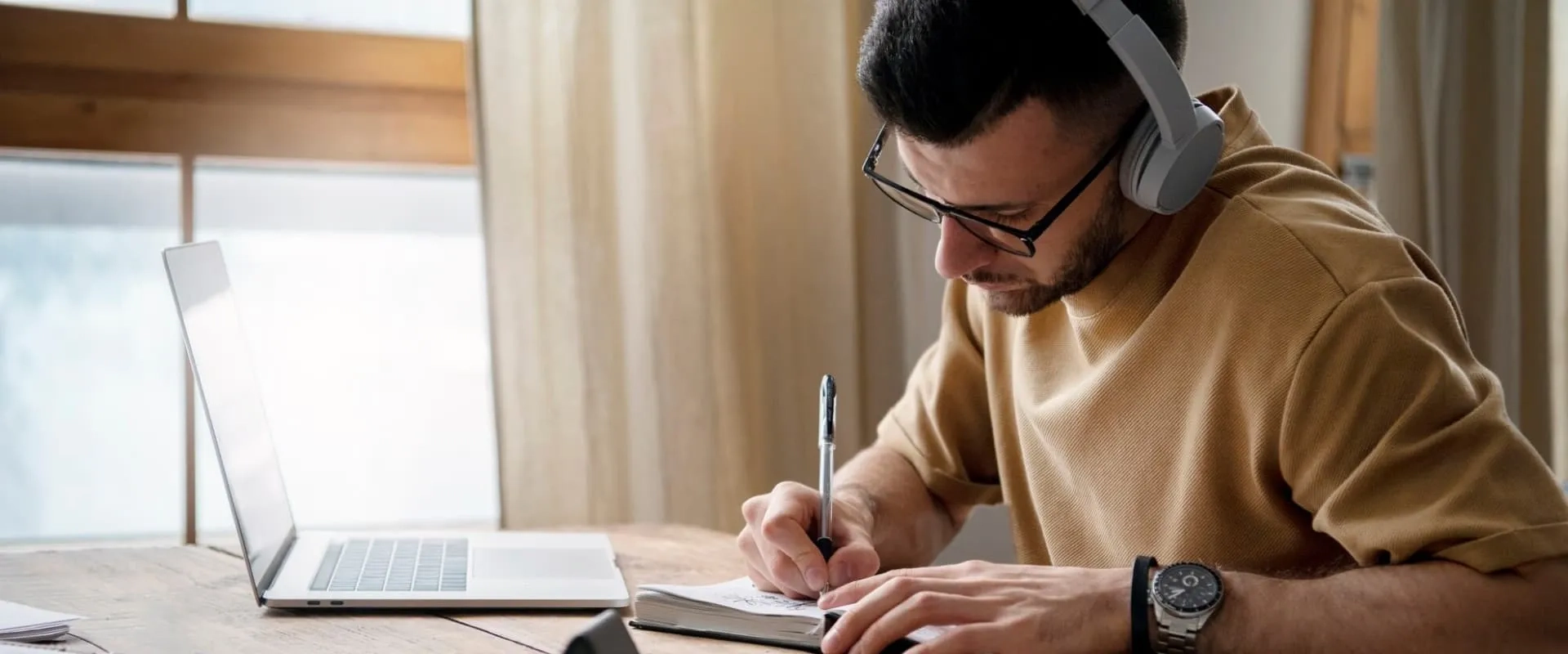 Joven estudiando en casa con laptop y audífonos, tomando notas para su educación en línea o aprendizaje a distancia.