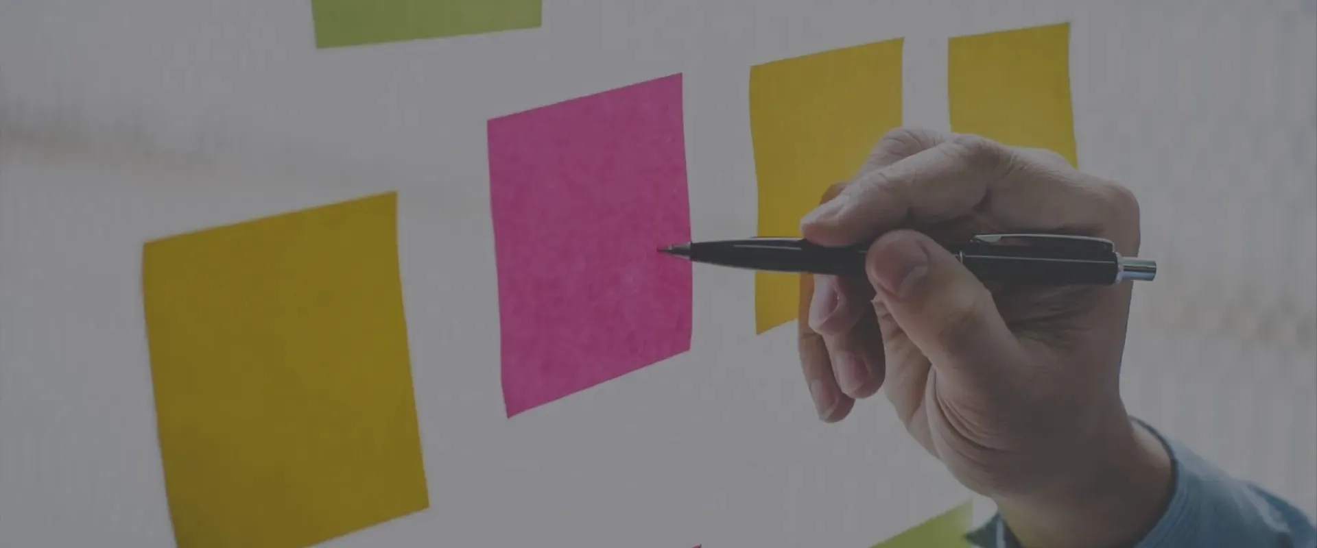 Una mano sostiene un bolígrafo negro mientras escribe en una nota adhesiva de color rosa, que está colocada sobre un fondo de notas adhesivas amarillas y verdes. 