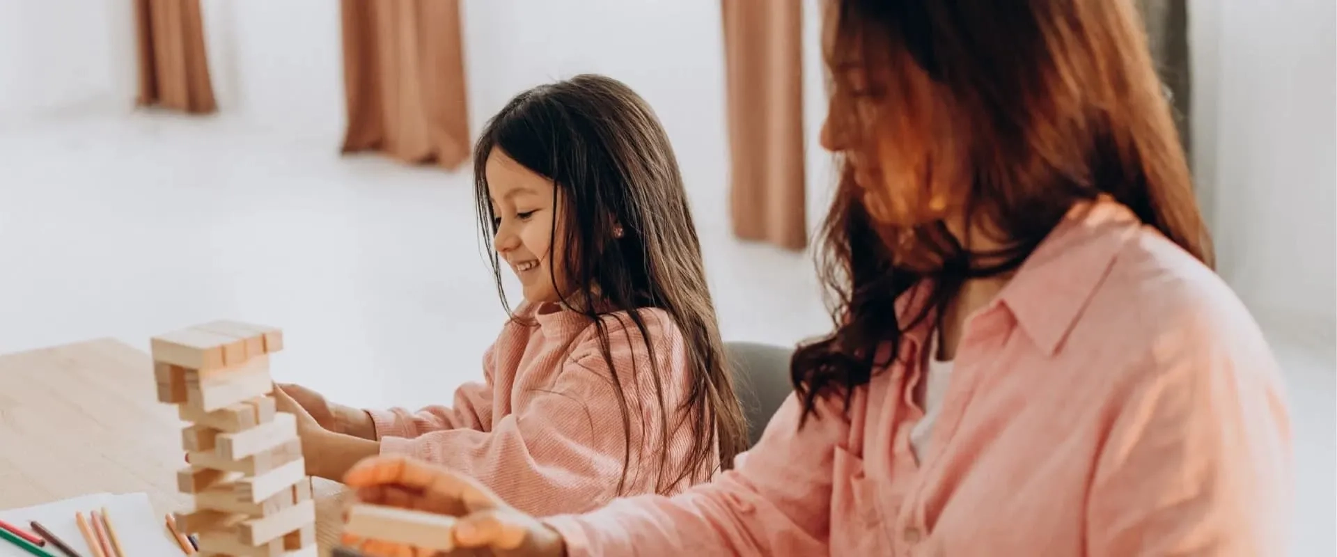 Una niña y una mujer están sentadas en una mesa, jugando con bloques de madera. La niña sonríe mientras juega, y la mujer la observa con atención.