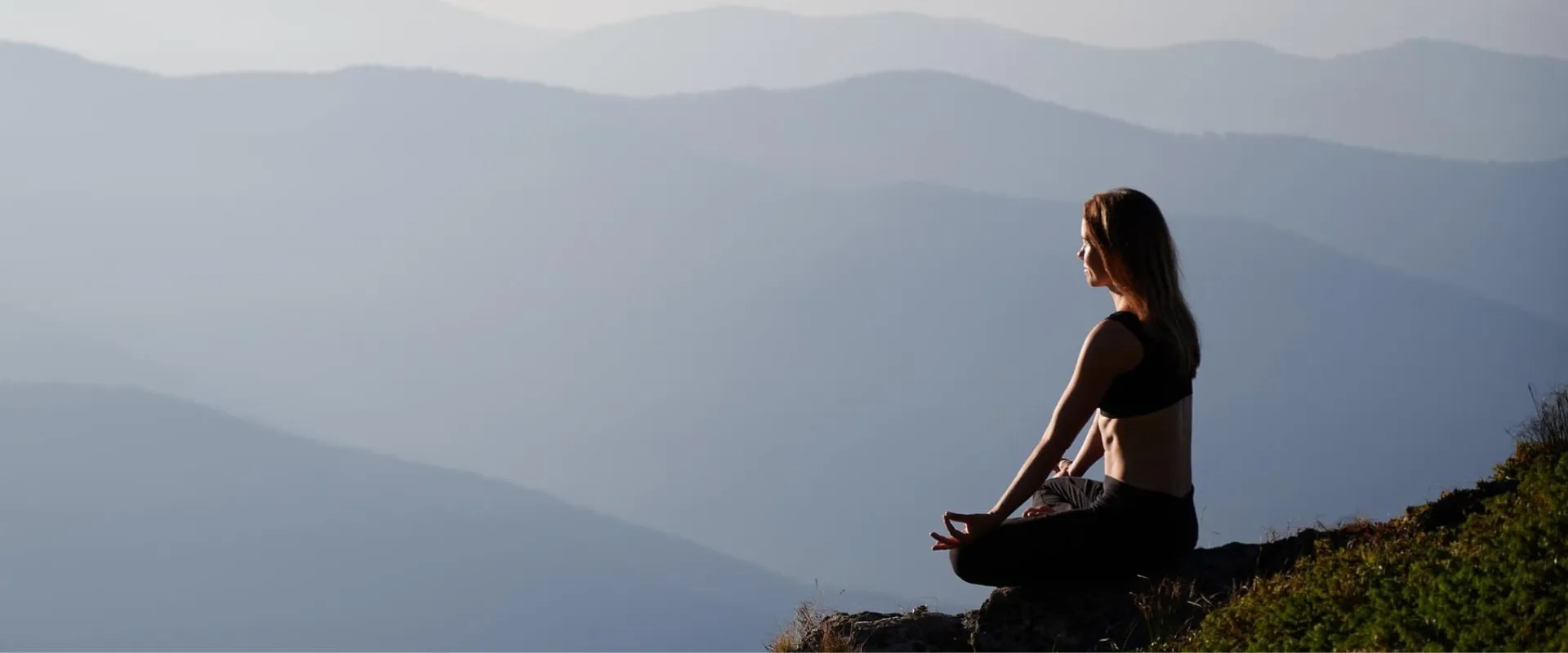 Una mujer está sentada en una roca en la cima de una montaña, meditando en una posición de loto. Viste un top negro y su cabello cae suelto. 