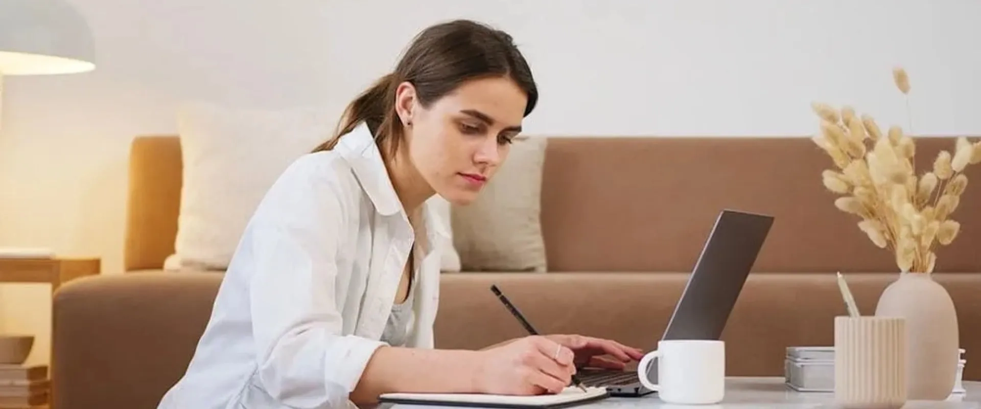 Una mujer está sentada en el suelo de una sala de estar moderna, escribiendo en un cuaderno mientras trabaja en una computadora portátil. Lleva una camisa blanca y parece concentrada en su tarea.