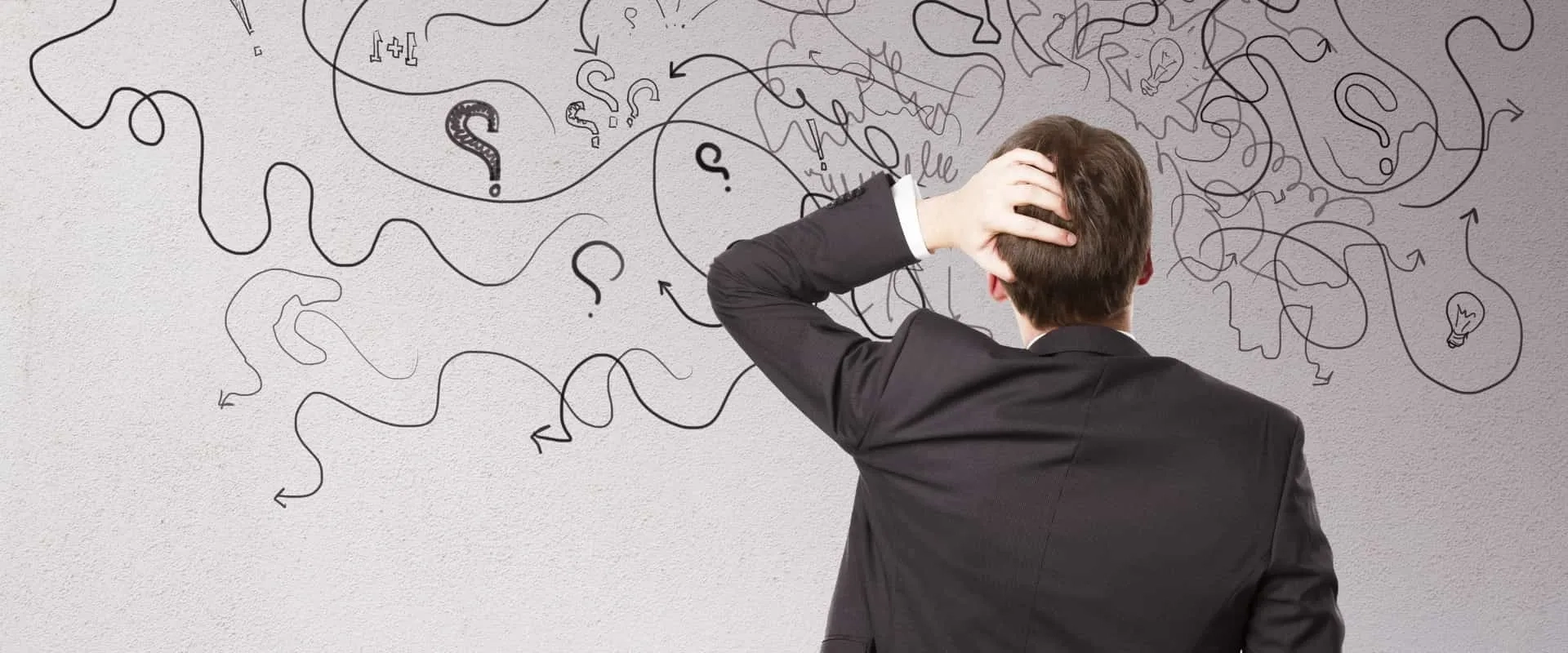 Un hombre de espaldas, vestido con un traje, se rasca la cabeza mientras observa un mural con garabatos y signos de interrogación. 