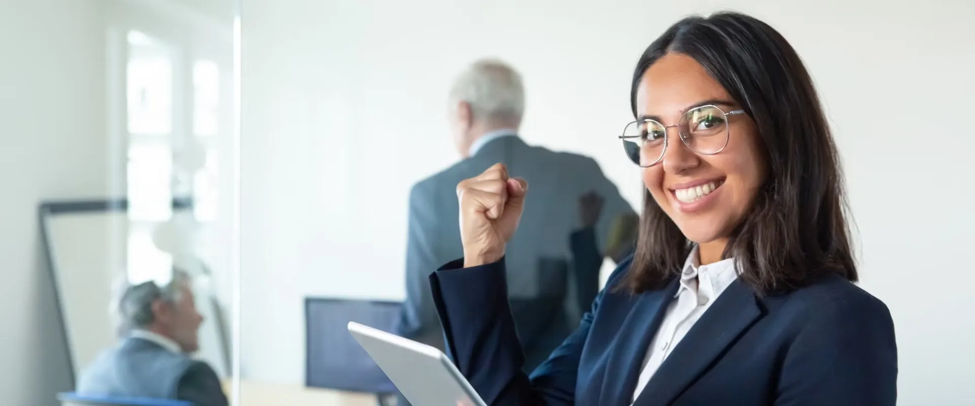 Una mujer sonriente en un entorno de oficina, vistiendo un traje formal y gafas. Sostiene una tablet con una mano levantada en señal de triunfo. 